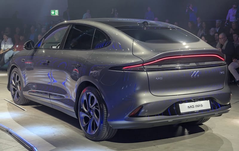 SUV cupê elétrico MG IM6 cinza visto de trás em uma apresentação, com a luz de freio em faixa vermelha contínua e público ao fundo