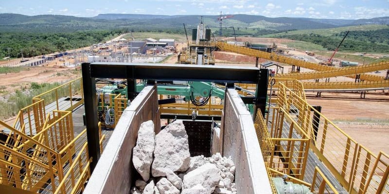 Blocos de rocha em uma caçamba de planta de beneficiamento mineral, com esteiras amarelas e a área da mina ao fundo
