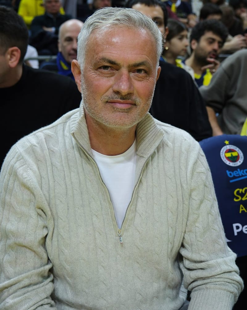 José Mourinho, de cabelo grisalho e barba rala, com suéter bege de zíper e camiseta branca, sentado em arquibancada lotada
