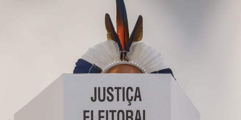 Pessoa com cocar de penas azuis e laranja aparece atrás de uma cabine de votação com a inscrição Justiça Eleitoral