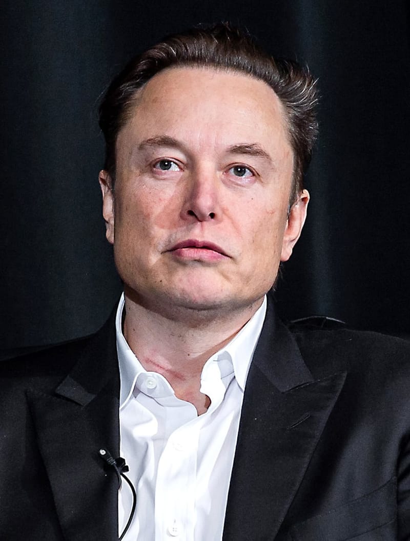 Elon Musk, de terno preto e camisa branca, olha de frente diante de um fundo escuro