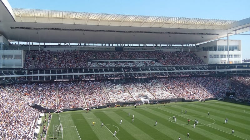Interior da Neo Química Arena em dia de jogo, com as arquibancadas lotadas, a cobertura branca e o gramado sob céu azul