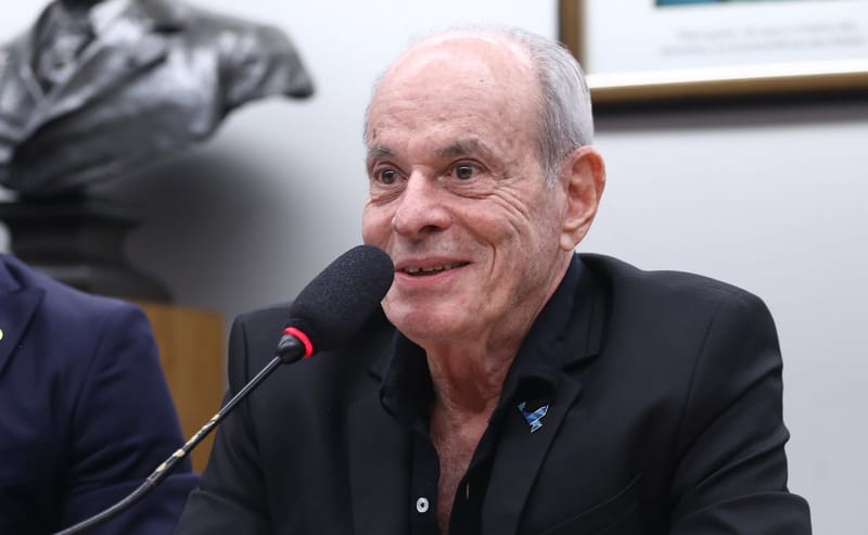 Ney Matogrosso, de cabelos grisalhos e roupa preta, sorri diante de um microfone durante audiência pública