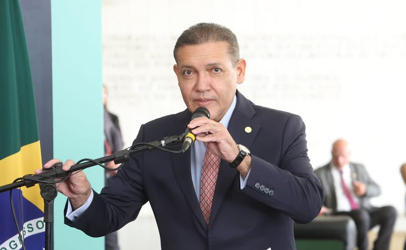 Kassio Nunes Marques, de terno azul-marinho e gravata vermelha, fala ao microfone ao lado de uma bandeira do Brasil