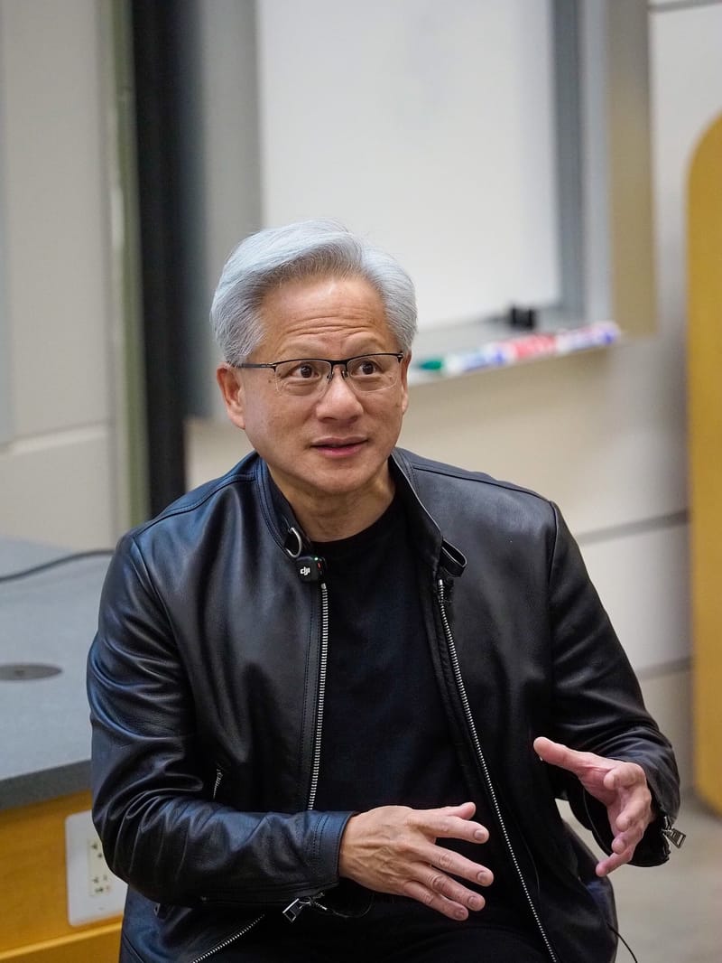 Jensen Huang, de óculos e jaqueta de couro preta, gesticula enquanto fala sentado numa sala de aula