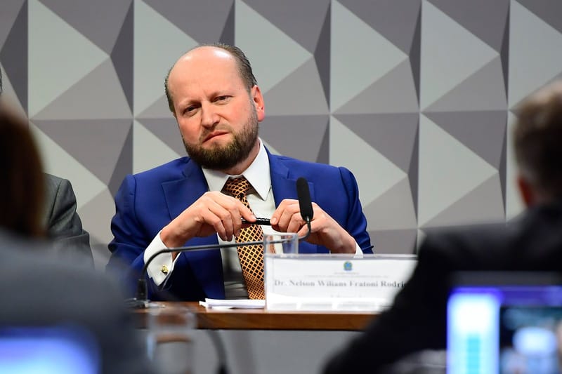 Nelson Wilians, de barba, terno azul e gravata laranja, sentado à mesa da CPMI diante de microfone e placa com seu nome