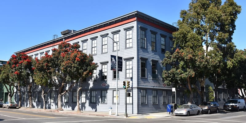 Prédio de três andares de madeira cinza com detalhes vermelhos, numa esquina de San Francisco, cercado por árvores floridas