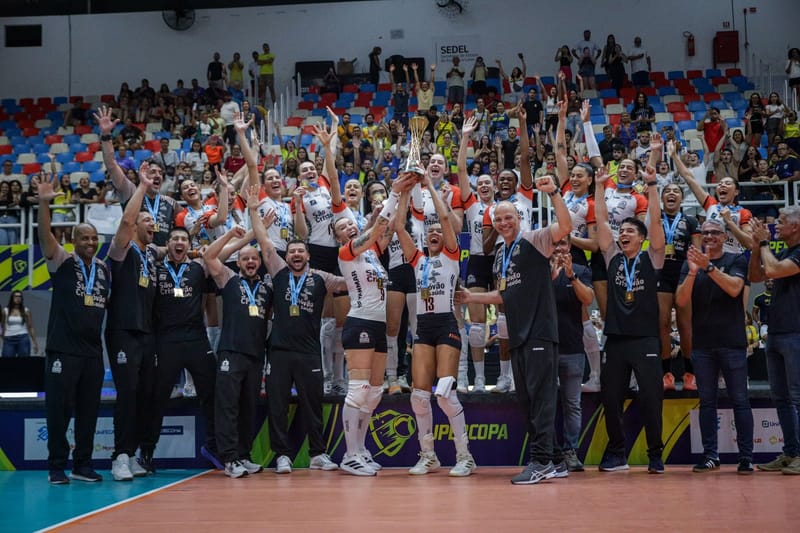 Jogadoras do Osasco São Cristóvão Saúde levantam o troféu da Supercopa no pódio, cercadas pela comissão técnica de braços erguidos e pela torcida na arquibancada