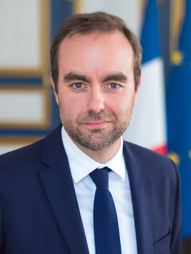 Retrato de Sébastien Lecornu, de terno azul-marinho e gravata, diante das bandeiras da França e da União Europeia