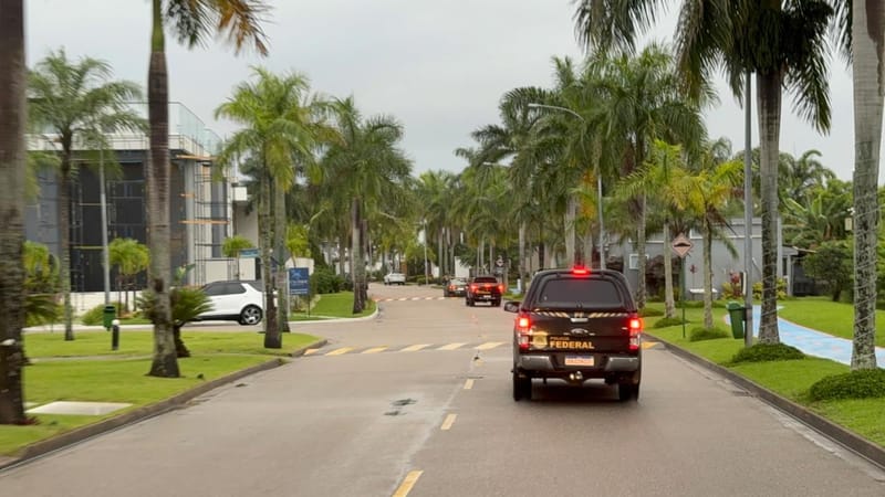 Três viaturas escuras da Polícia Federal descem uma rua de condomínio com palmeiras dos dois lados