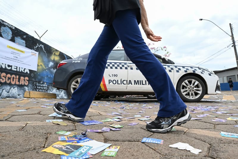 Pessoa caminha sobre santinhos no chão, ao lado de viatura da Polícia Militar, diante de local de votação no Distrito Federal