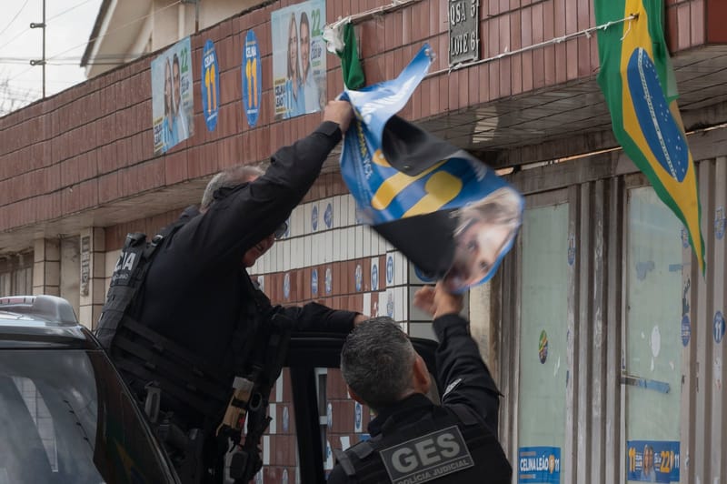 Dois policiais de preto arrancam cartaz de candidato da fachada de um imóvel, com outros cartazes e uma bandeira do Brasil