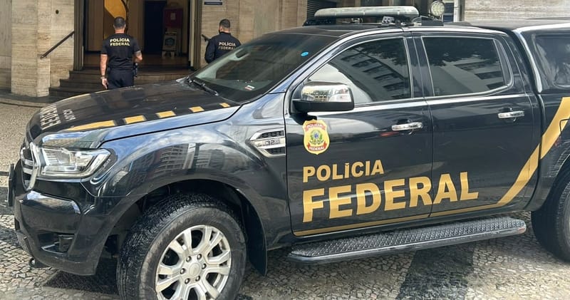 Viatura preta da Polícia Federal estacionada diante de um prédio, com dois agentes na entrada