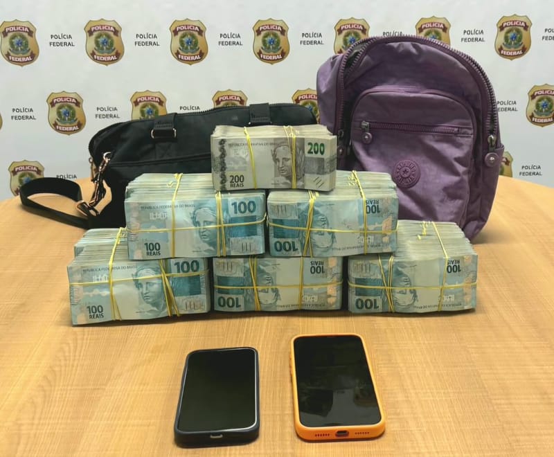 Maços de notas de 100 e 200 reais, duas bolsas e dois celulares sobre mesa diante de painel da Polícia Federal