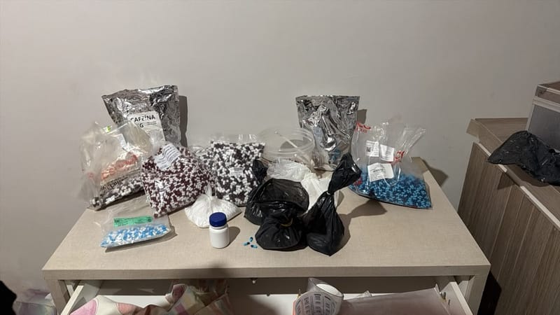 Sobre uma mesa bege, sacos com cápsulas bicolores, embalagens metálicas de insumos e um pote branco apreendidos pela PF