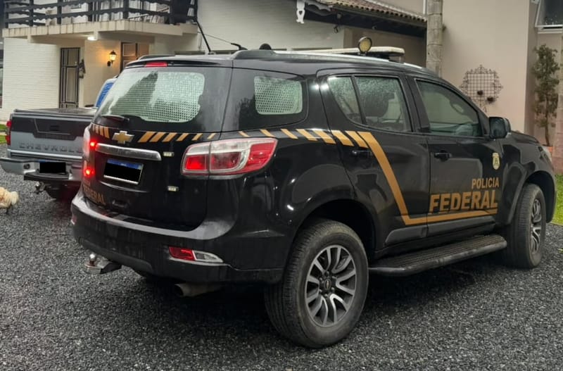 Viatura preta da Polícia Federal estacionada no cascalho diante de uma casa, ao lado de uma caminhonete