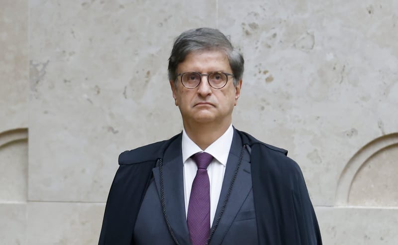 Paulo Gonet, de óculos e beca escura, olha de frente para a câmera diante de uma parede de pedra clara