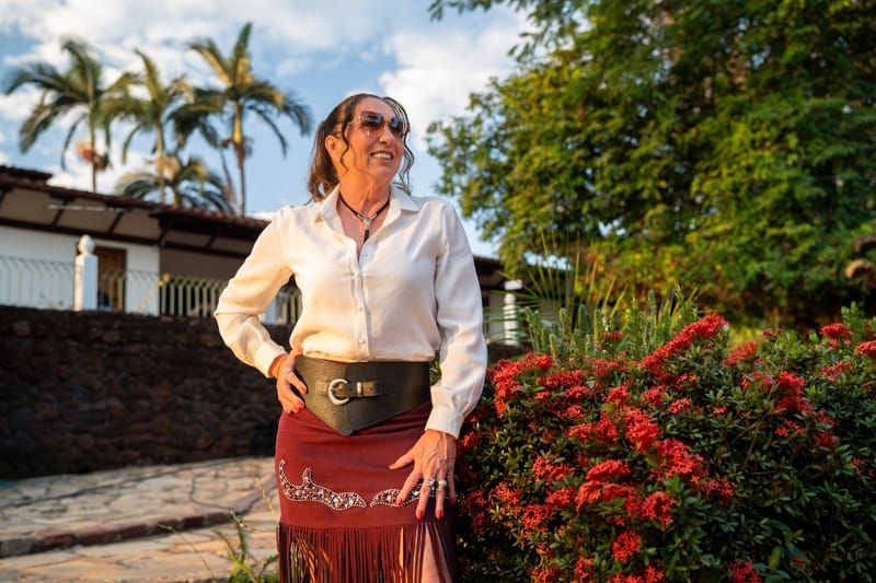 Margareth Serrão, de óculos escuros, blusa branca e saia vinho, sorri ao lado de arbustos floridos, com palmeiras ao fundo