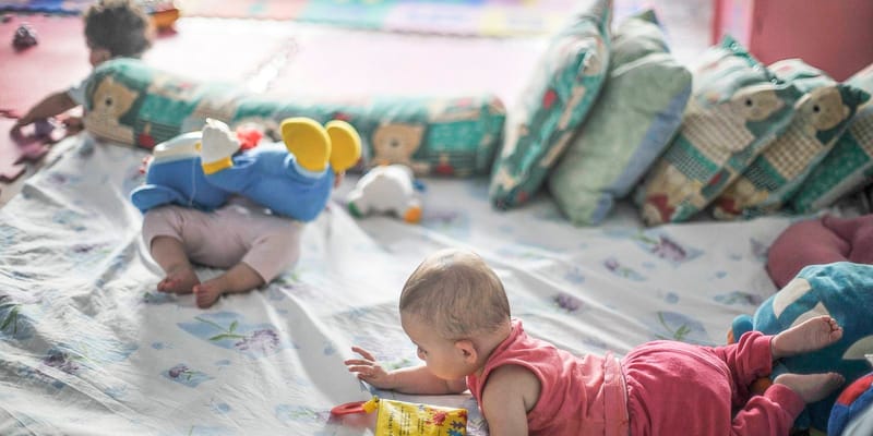 Dois bebês brincam deitados sobre colchonetes e almofadas estampadas, em um ambiente infantil colorido