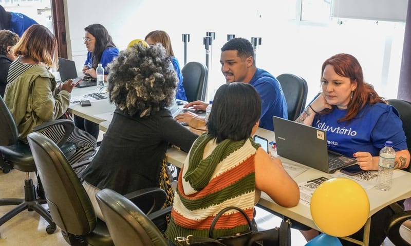Voluntários de camiseta azul atendem refugiados e migrantes em mesas com notebooks durante a ação de empregabilidade no centro de São Paulo