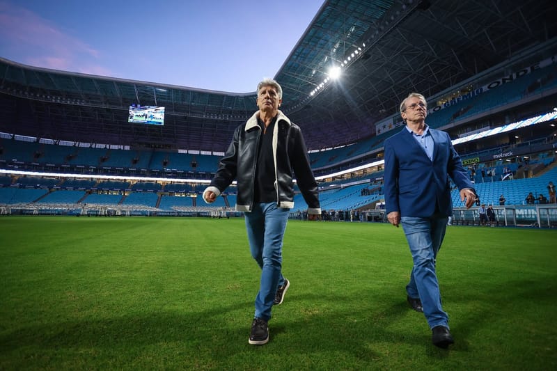Renato Portaluppi, de jaqueta preta, caminha pelo gramado da Arena do Grêmio ao lado de um dirigente de blazer azul