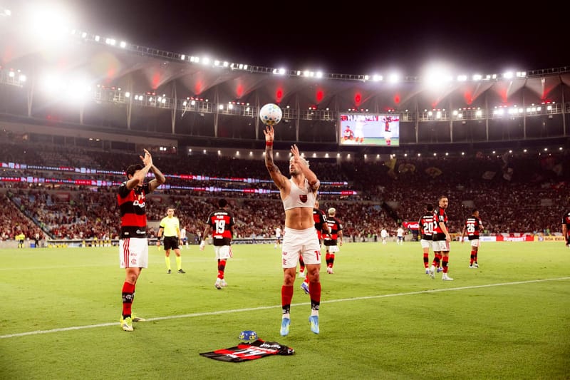 Jogador do Flamengo, sem camisa e com os braços erguidos segurando a bola, diante da torcida no estádio iluminado, com um companheiro aplaudindo ao lado