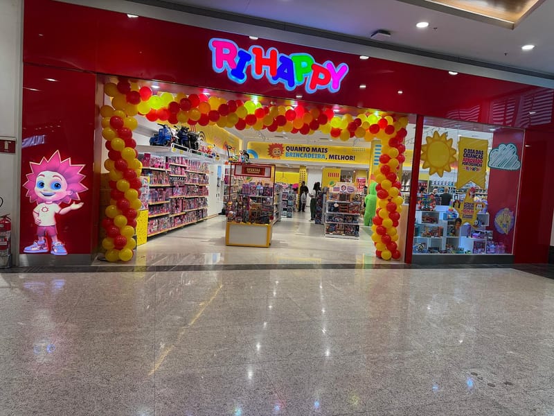 Fachada vermelha de uma loja da Ri Happy em shopping, com letreiro colorido, arco de balões e a mascote ao lado da entrada