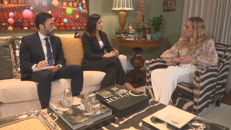Roberta Luchsinger, de cabelo loiro e blusa estampada, sentada em uma poltrona listrada, conversa com dois jornalistas sentados em um sofá, em uma sala decorada
