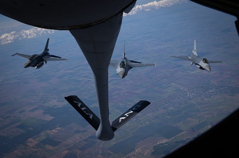 Três caças F-16 da Força Aérea Romena voam em formação atrás do bocal de reabastecimento de um avião-tanque, sobre campos agrícolas