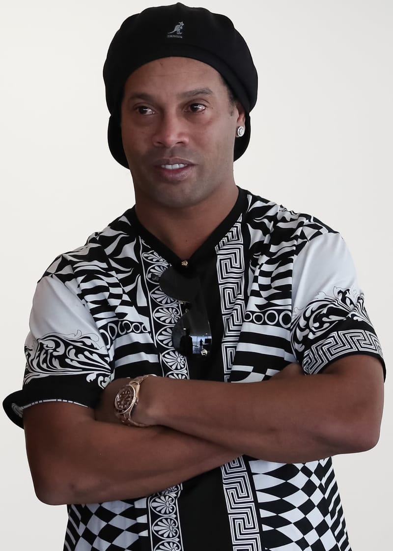 Ronaldinho Gaúcho, de boina preta e camisa estampada em preto e branco, olha para o lado com os braços cruzados