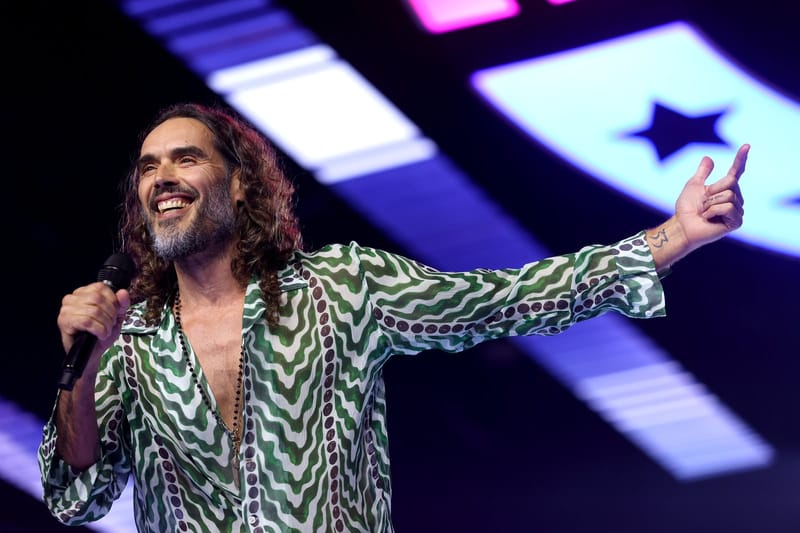 Russell Brand, de cabelos longos e barba, sorri com um microfone na mão e o braço aberto num palco com luzes coloridas