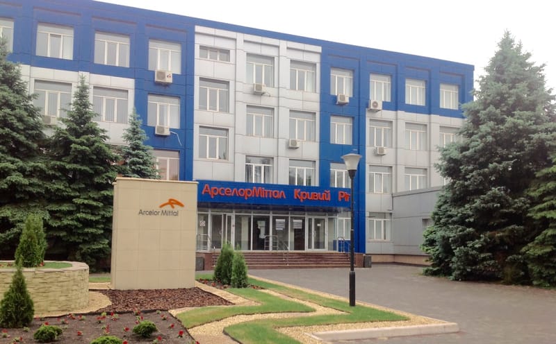 Prédio administrativo azul e cinza da ArcelorMittal Kryvyi Rih, com o nome da empresa em letreiro vermelho na fachada e pinheiros ao redor