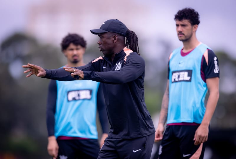 Clarence Seedorf, de boné e agasalho preto do Corinthians, gesticula com os braços estendidos diante de dois jogadores de colete azul no CT