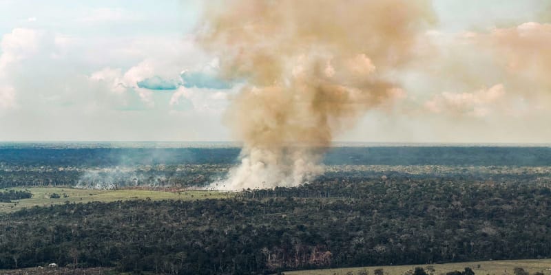 Coluna de fumaça de uma queimada se ergue sobre a mata em vista aérea, com pasto à esquerda e céu parcialmente nublado