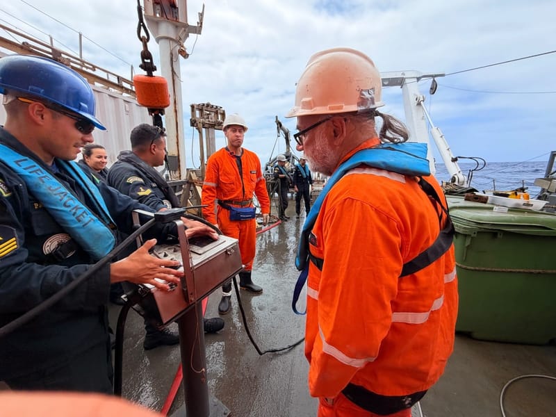 Pesquisadores de macacão laranja e militares da Marinha operam equipamento no convés do navio de pesquisa, com o mar ao fundo