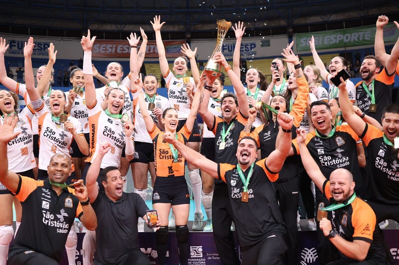 Jogadoras e comissão técnica do Osasco São Cristóvão Saúde, de uniformes laranja, branco e preto, erguem o troféu da Supercopa e as medalhas na quadra