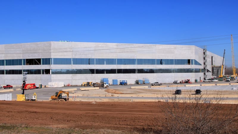 Fachada de concreto e vidro de grande fábrica em obras, com guindastes e máquinas no terreno, sob céu azul