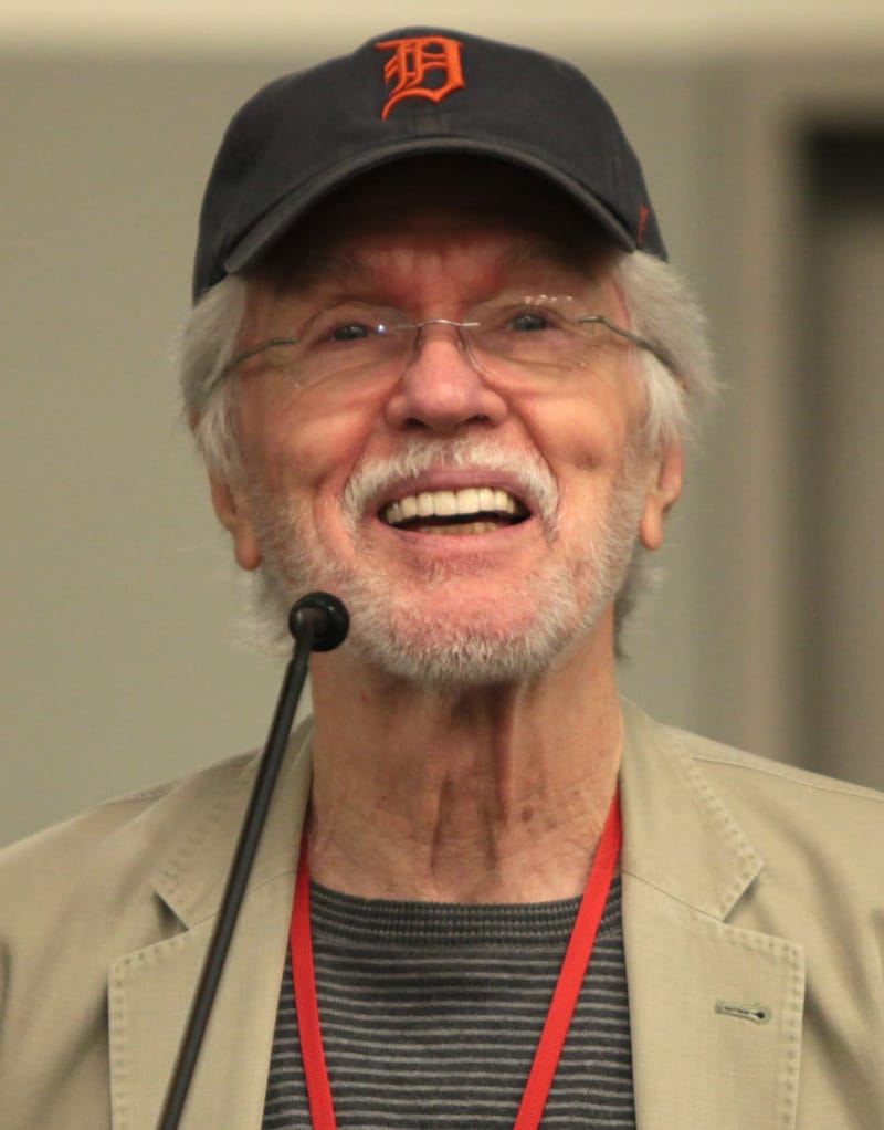 Tom Skerritt, de boné preto, óculos e barba branca, sorri diante de um microfone