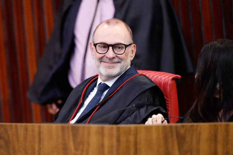 Ministro Ricardo Villas Bôas Cueva, de óculos de armação escura e barba grisalha, sorri sentado na bancada do plenário do TSE, de toga com cordão vermelho