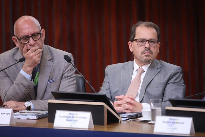 Floriano de Azevedo Marques, de óculos e terno cinza, sentado à mesa do TSE atrás da placa com seu nome