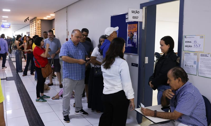 Eleitores fazem fila no corredor de um local de votação, diante da porta da seção 0357, onde um mesário confere documentos sentado numa carteira