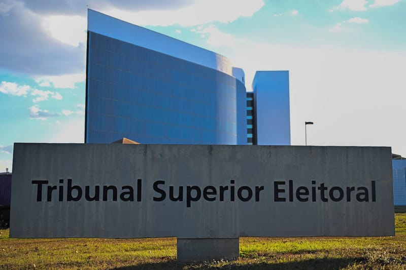 Placa com o nome Tribunal Superior Eleitoral em frente ao prédio de vidro azul do TSE, em Brasília, sob céu com nuvens