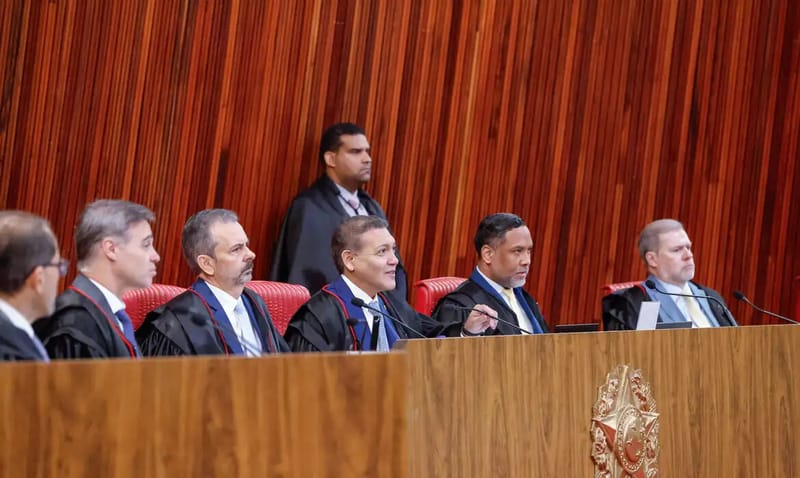 Ministros do TSE, de toga preta, sentados lado a lado na bancada de madeira do plenário, com o painel de madeira ao fundo