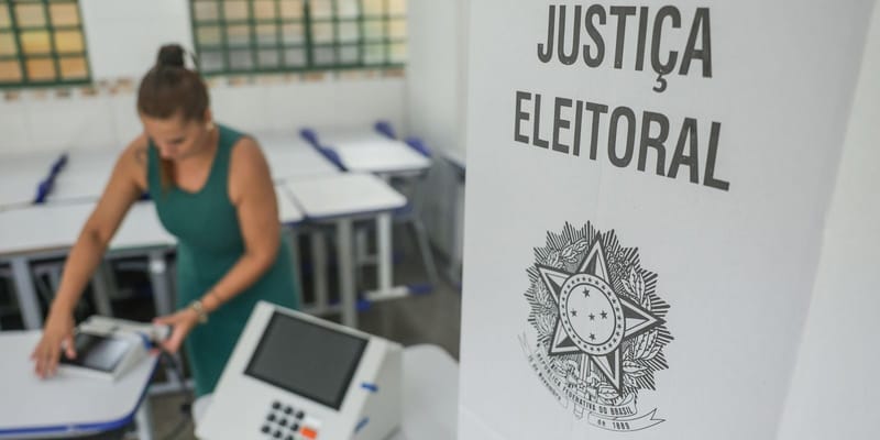 Mesária prepara a urna eletrônica numa sala de aula; à frente, a cabine com o brasão e os dizeres Justiça Eleitoral