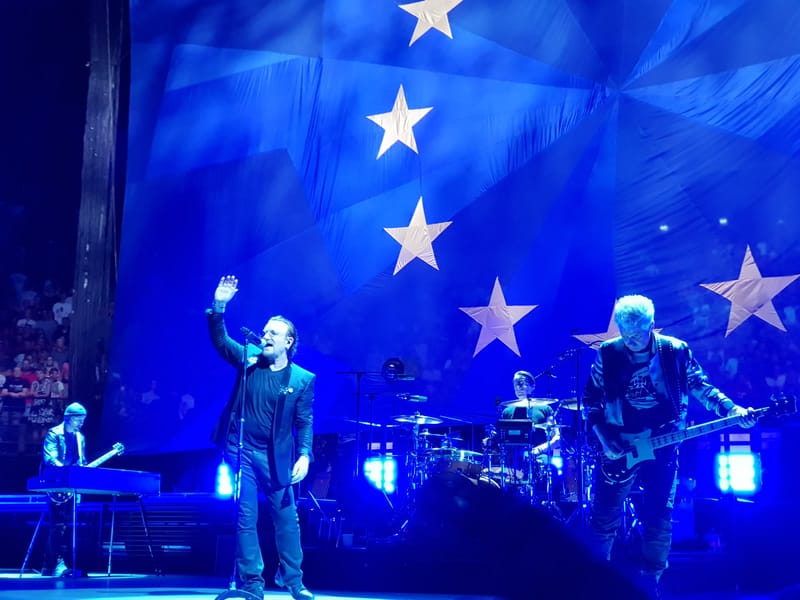 Bono acena ao público ao lado dos integrantes do U2 durante show, sob luz azul