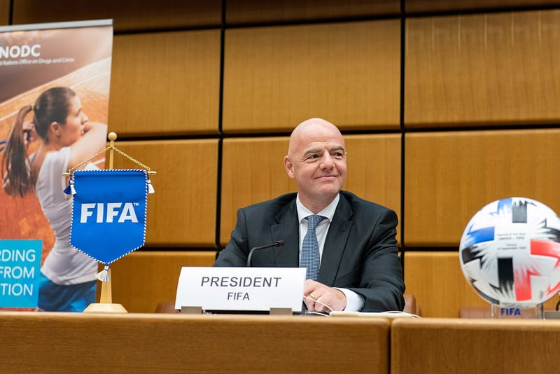 Gianni Infantino, de terno escuro e gravata azul, sorri atrás de placa com a inscrição President FIFA