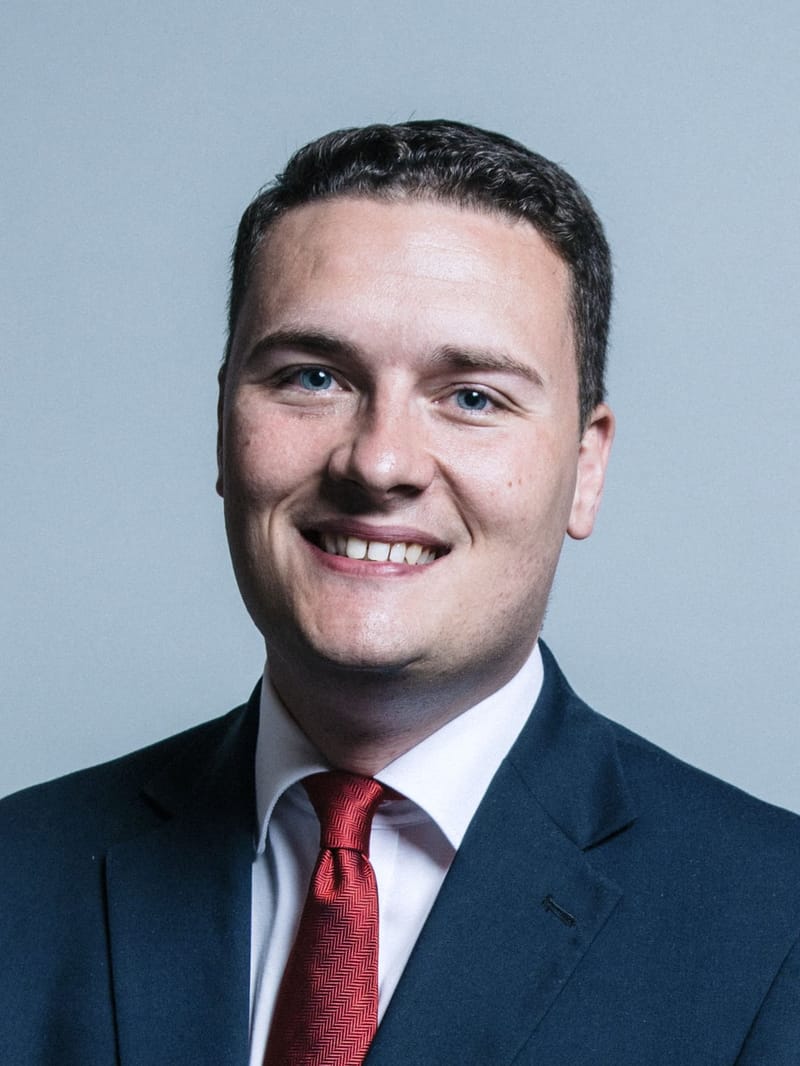 Retrato oficial de Wes Streeting, de terno azul-escuro e gravata vermelha, sorrindo diante de fundo cinza