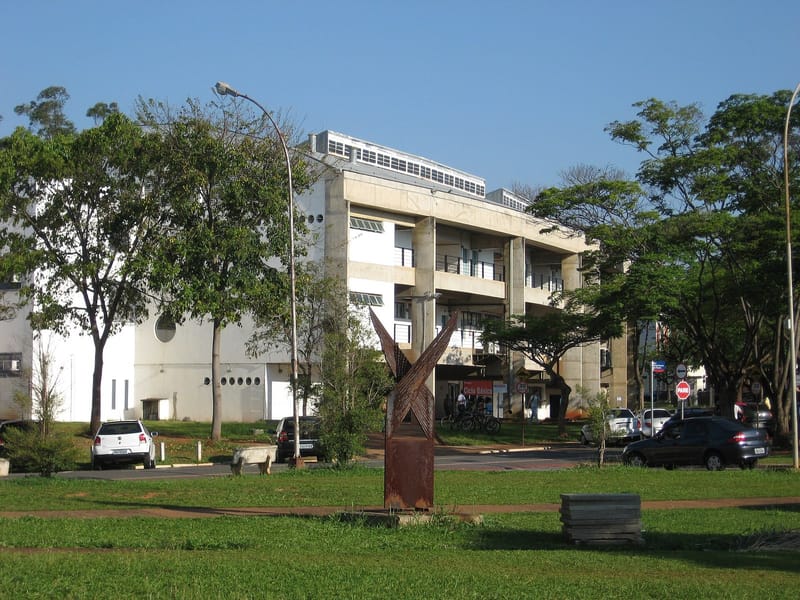 Prédio claro de concreto com três andares, o Pavilhão Básico da Unicamp, visto atrás de árvores e de uma escultura de metal enferrujado, sob céu azul