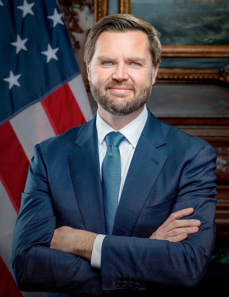 JD Vance, de terno azul e gravata azul-clara, de braços cruzados diante da bandeira dos Estados Unidos