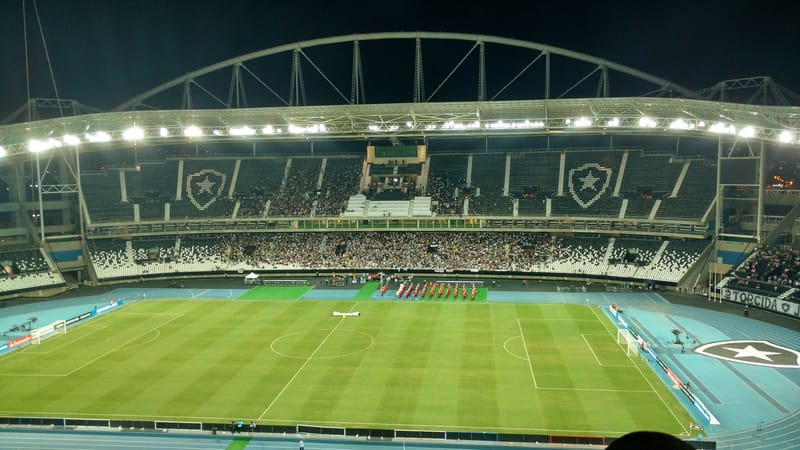 Vista noturna do gramado e das arquibancadas do Nilton Santos, com a cobertura em arco iluminada e torcida nos setores